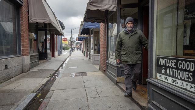 Grok Imagine scene: a man stepping out of a shop doorway onto a narrow sidewalk ahead